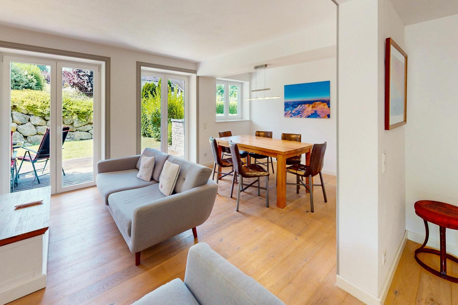 Wohn- und Essbereich mit Sofa, Tisch und Stühlen. Durchgänge zu einem Garten.