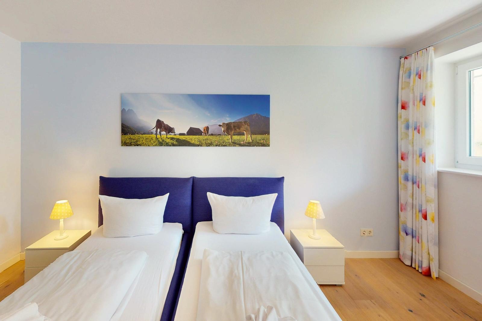 Zwei Betten mit weißen Laken, blauer Kopfteilwand und Fenster mit bunten Vorhängen.