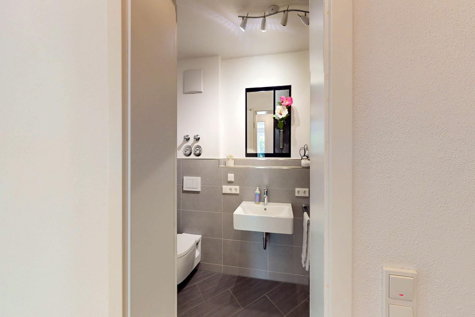 Badezimmer mit Waschbecken, Spiegel und Toilette. Graue Fliesen und weiße Wand.