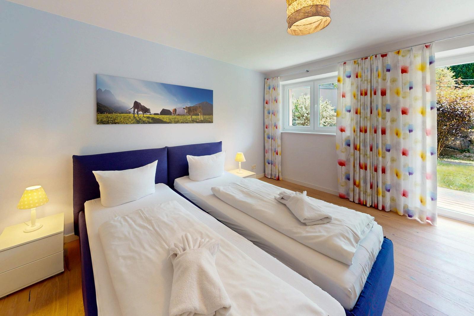 Zwei Einzelbetten mit weißen Laken, Fenster mit bunten Vorhängen und Wandbild mit Kühen.