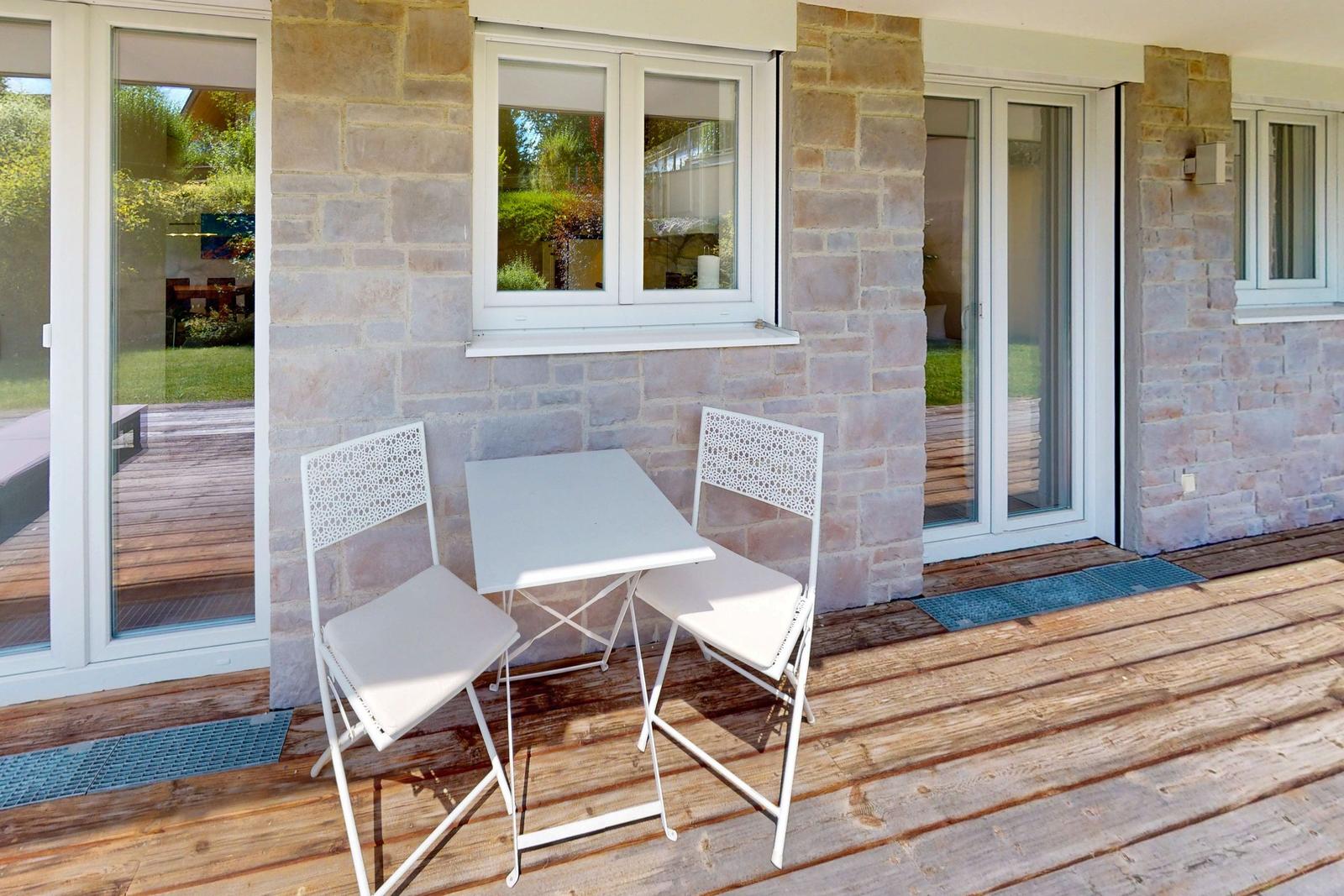 Terrasse mit weißem Tisch und zwei Stühlen vor Steinmauer und Glasfront.