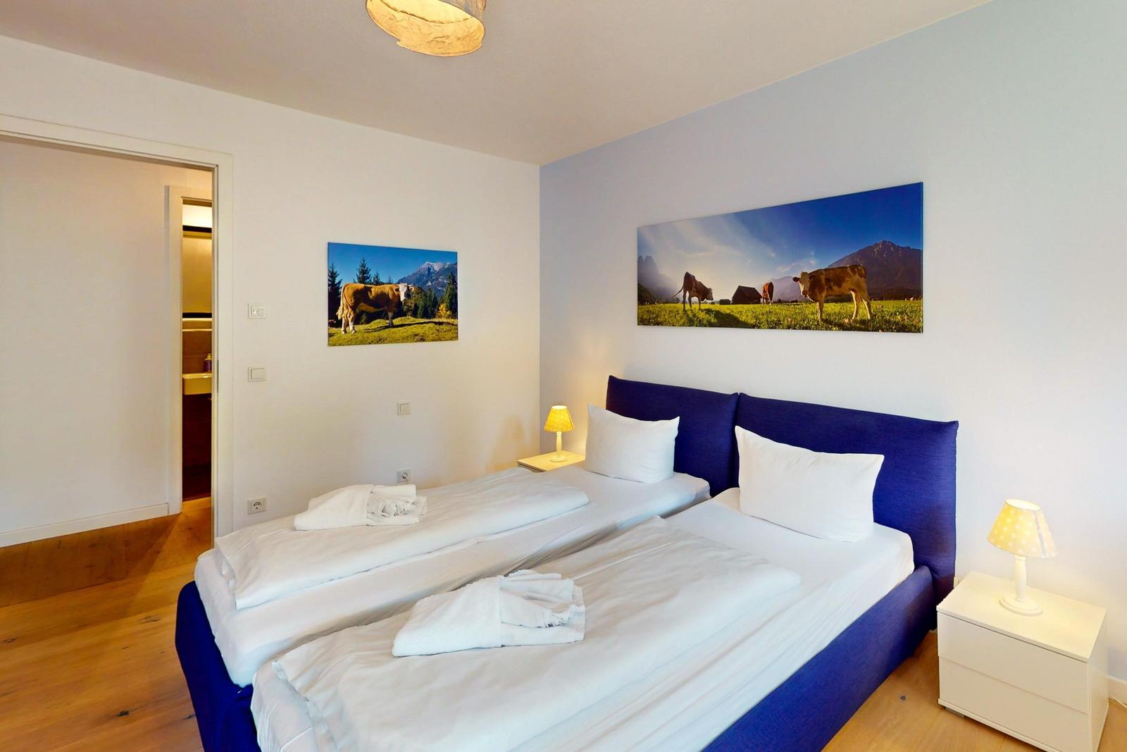 Zwei Betten mit weißen Bettwäsche und Kissen. Wandbilder mit Kühen und Bergen. Holzboden und Nachttische.