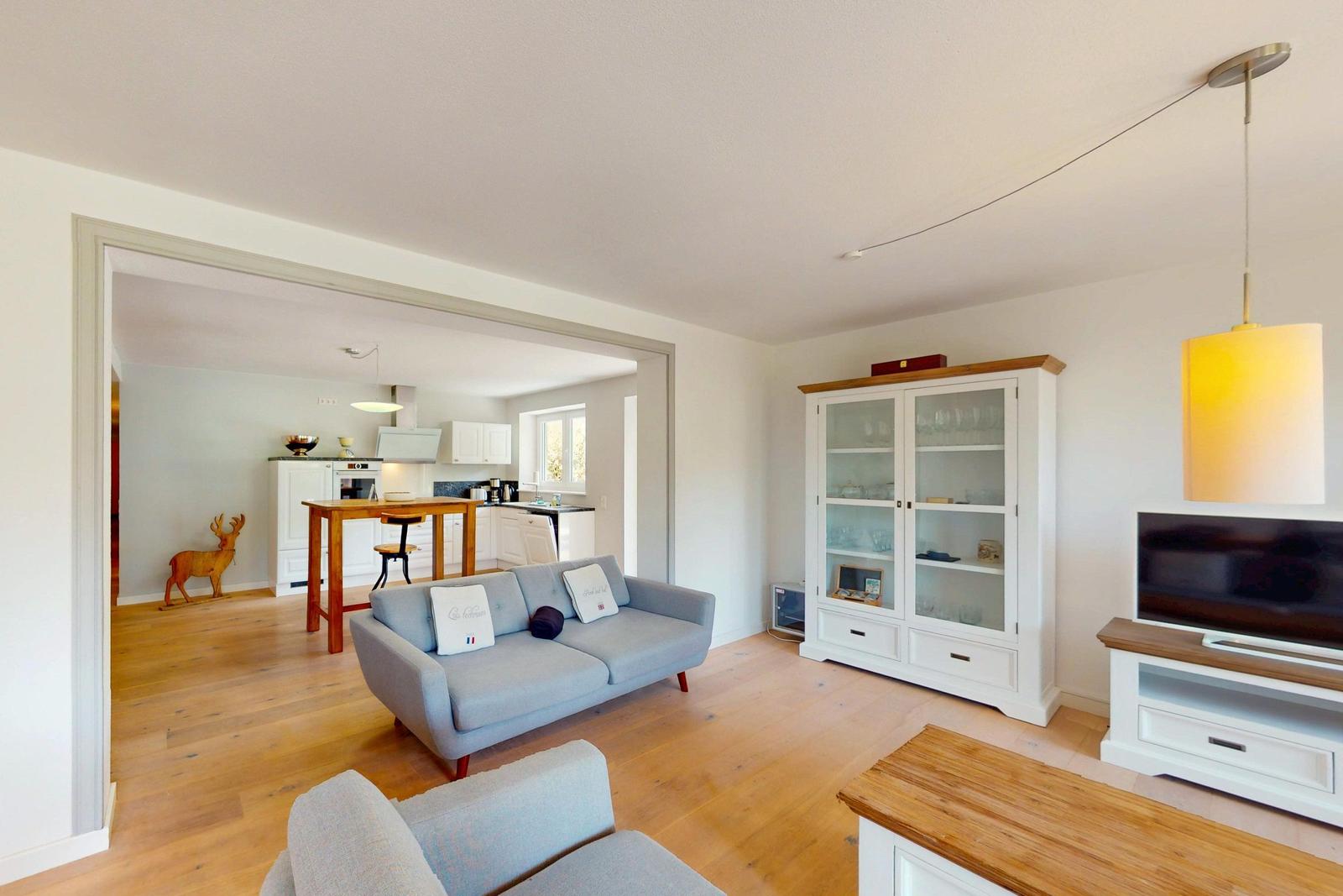 Wohnzimmer mit Sofa, TV und Glasvitrine. Küche im Hintergrund sichtbar.