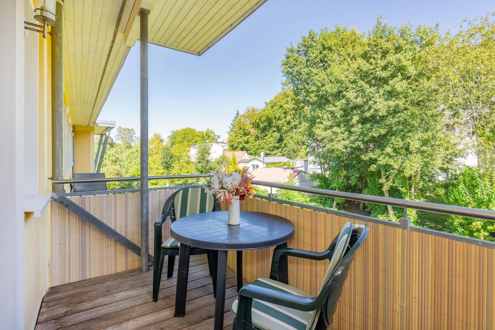 Terrasse mit Tisch und zwei Stühlen, Blick auf grüne Bäume und Häuser.