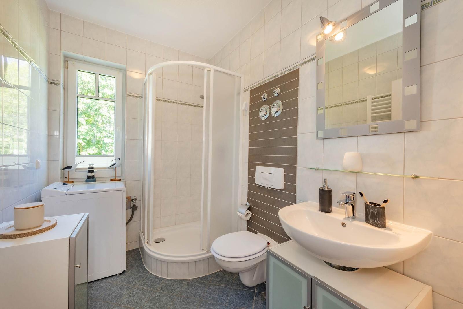 Badezimmer mit Dusche, Waschbecken und Toilette. Fenster mit Blick auf Grün.