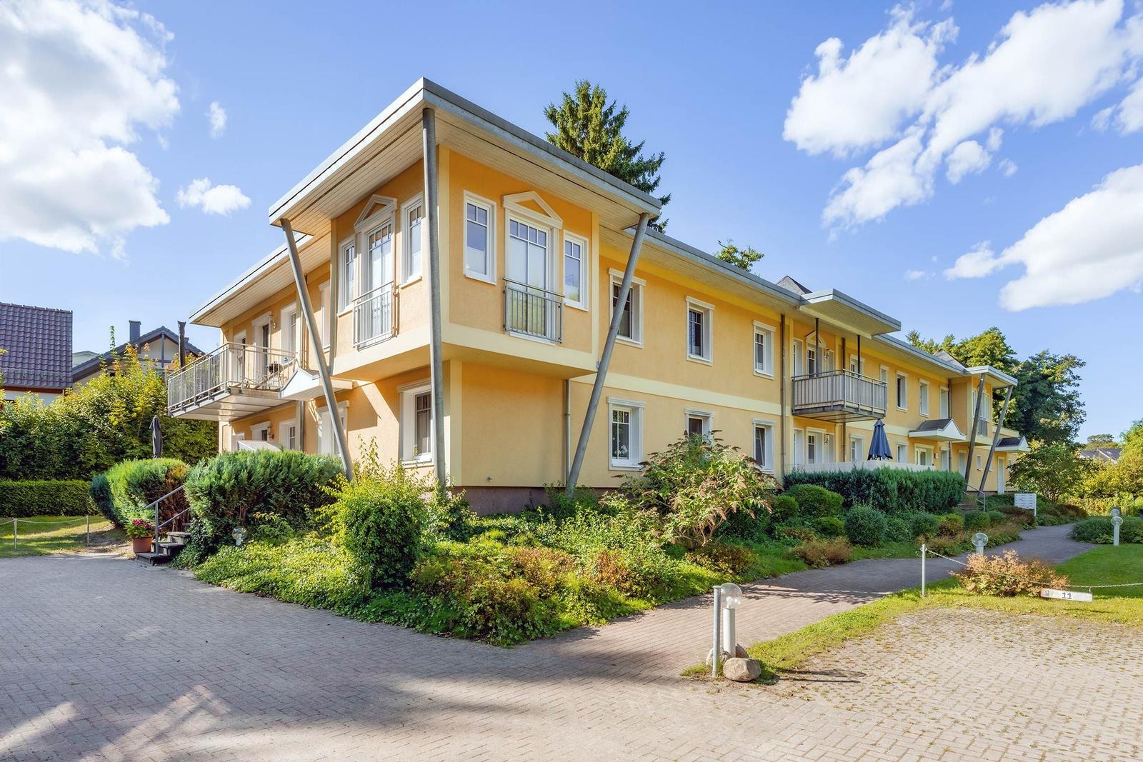 Zweistöckiges gelbes Haus mit Balkonen und gepflegtem Garten unter blauem Himmel.