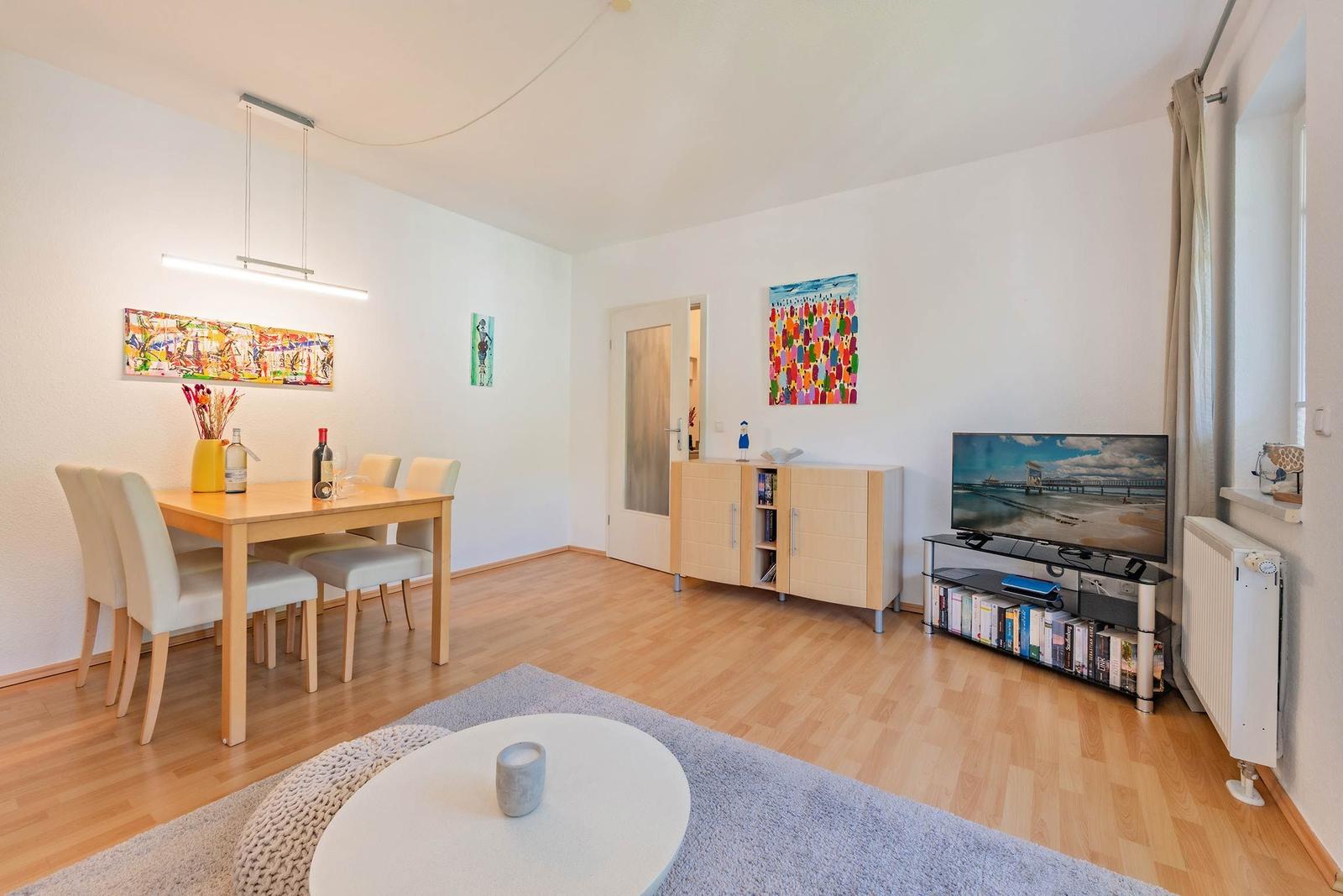 Wohn- und Essbereich mit Holzboden, TV, Tisch und Stühlen sowie Wandbildern.