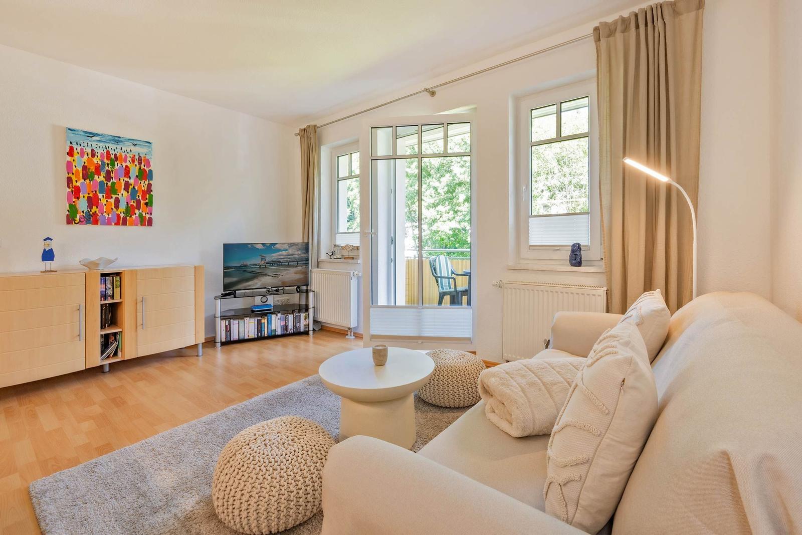 Wohnzimmer mit Sofa, Couchtisch und TV. Balkon mit Sitzgelegenheit. Fenster mit Vorhängen.