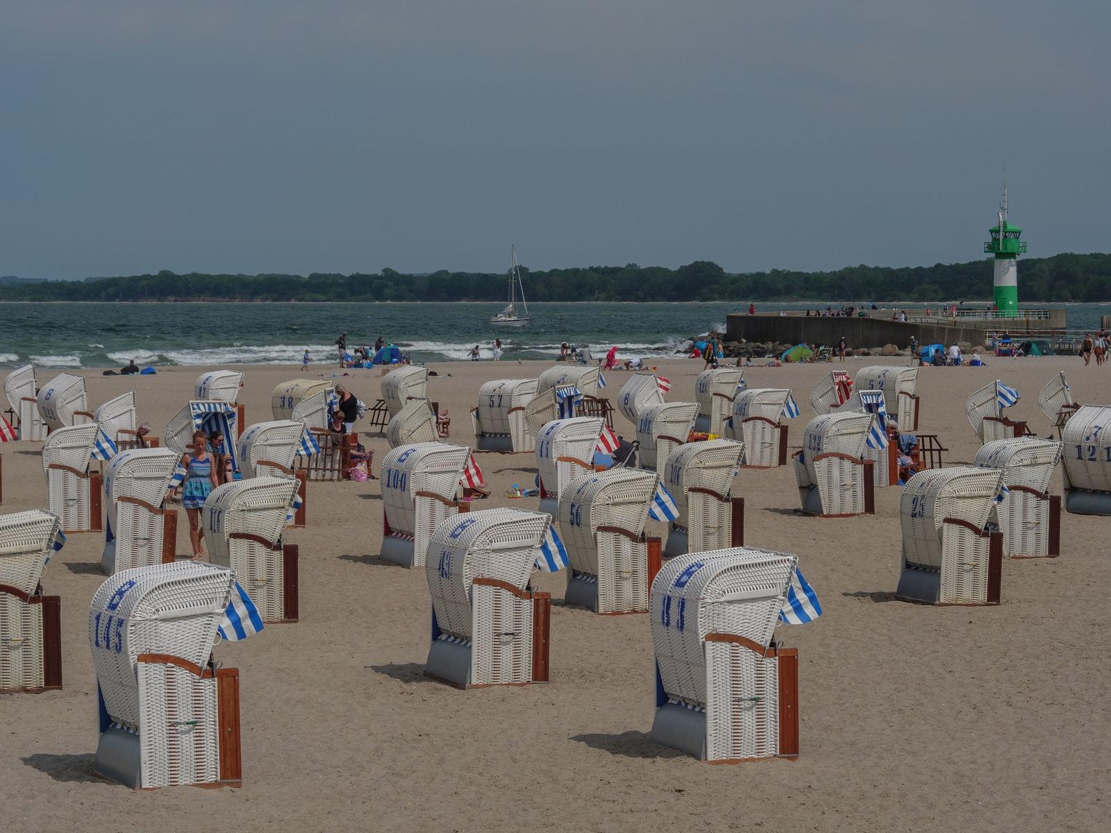 Strand mit Strandkörben, Leuchtturm und Segelyacht im Hintergrund.