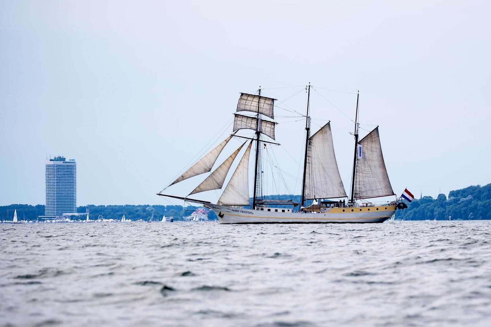 Segelschiff mit Segeln auf dem Wasser, im Hintergrund Gebäude und Bäume.