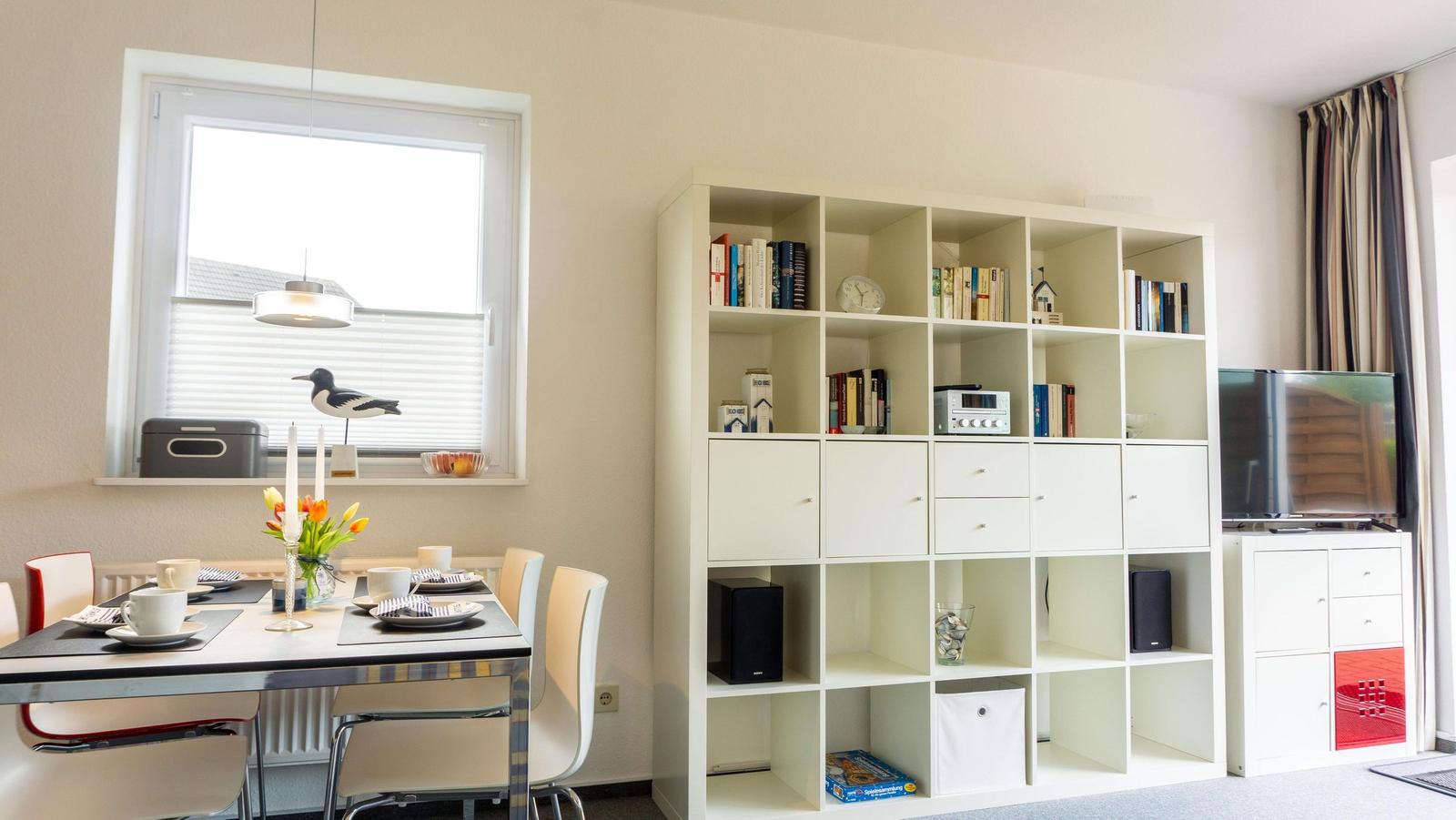 Essbereich mit Tisch, Stühlen und Bücherregal. Fenster mit Jalousie und Vogeldekoration.