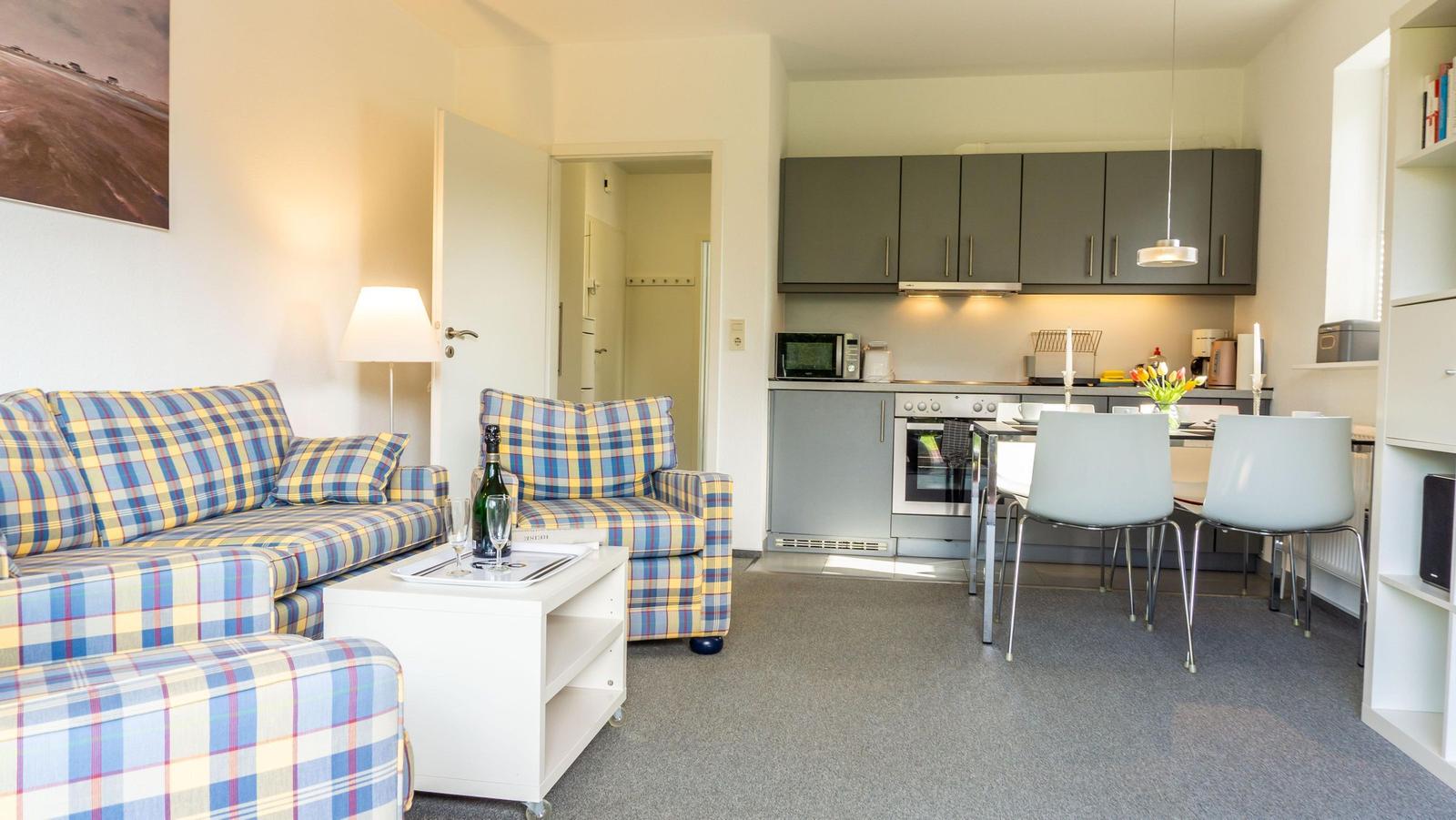 Wohnzimmer mit Plaidsofa, Küchenbereich mit Esstisch und grauen Schränken.