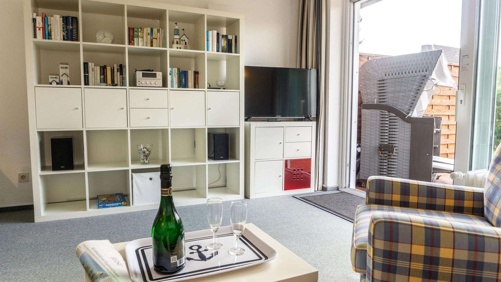 Wohnzimmer mit Bücherregal, TV und Sitzgelegenheit. Draußen liegt ein Wickerstuhl.