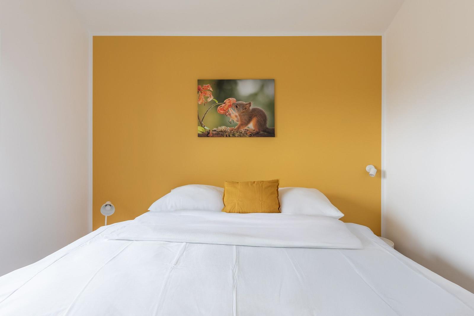 Schlafzimmer mit Bett, gelber Wand und Wandbild mit Eichhörnchen.