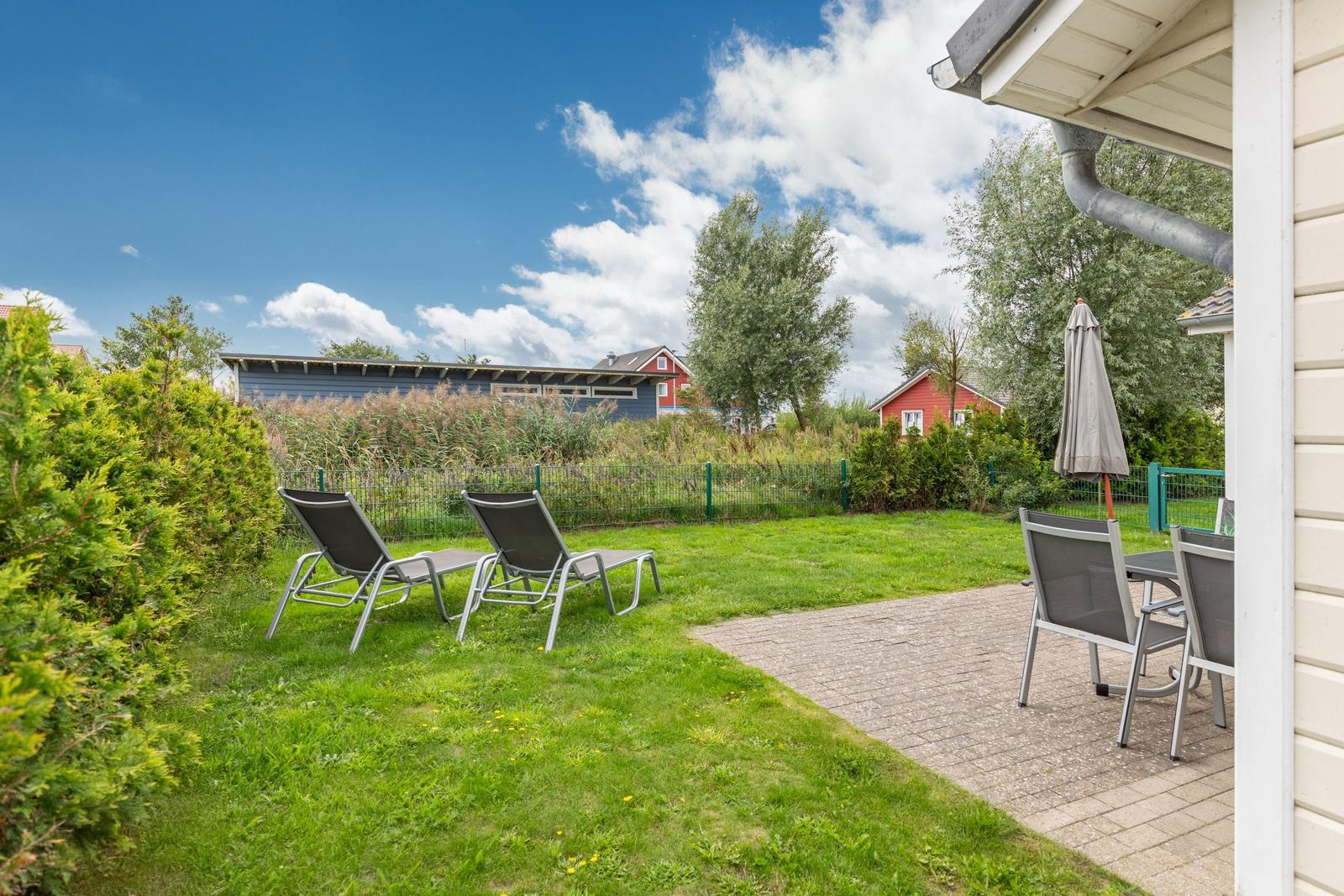 Garten mit Liegen und Tisch unter einem Schirm. Hinterhof mit Gras und Bäumen.