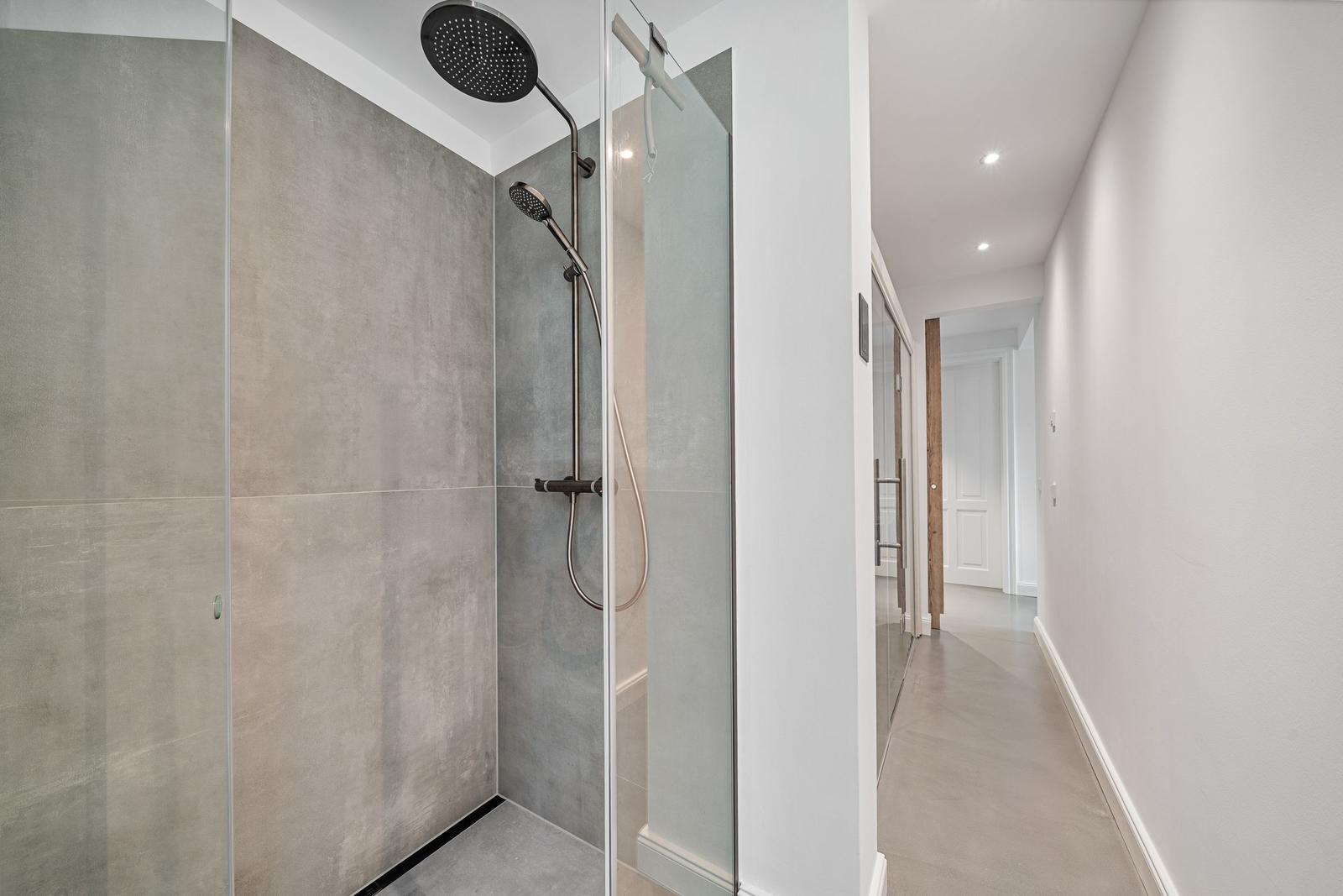 Duschkabine mit Glaswand und Duschbrause. Flur mit weißen Wänden und Boden.