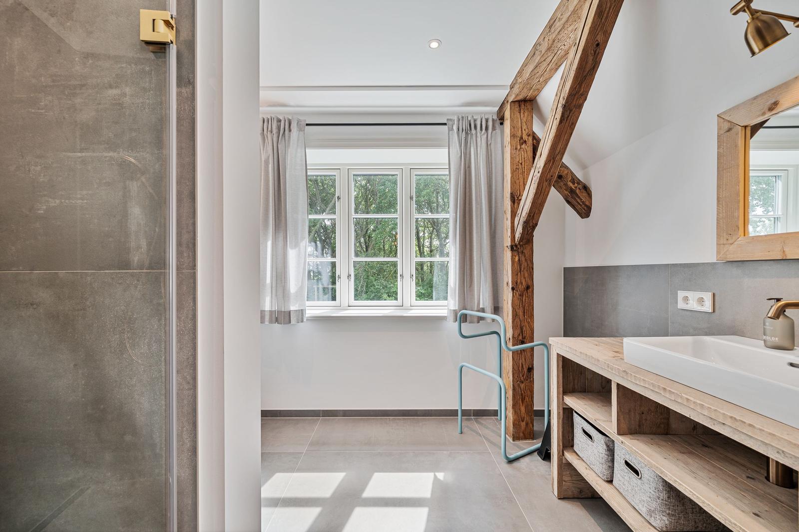 Badezimmer mit Dusche, Waschbecken und Holzrahmenspiegel. Holzbalken und Fenster mit Blick auf Bäume.