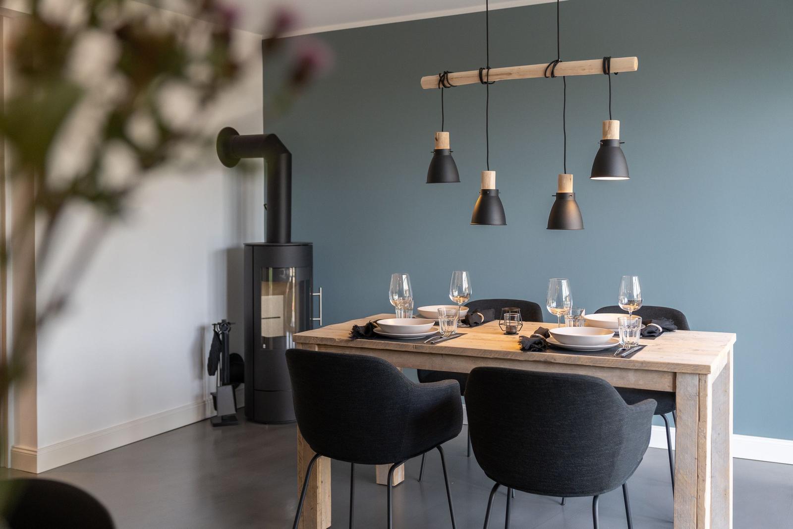 Esstisch mit sechs Stühlen und moderner Hängelampe. Im Hintergrund steht ein Holzofen.