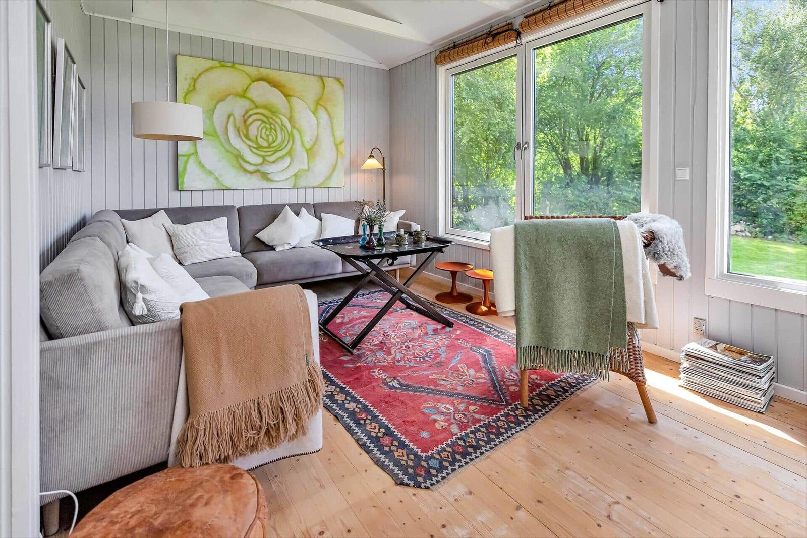 Wohnzimmer mit Sofa, Teppich und großen Fenstern. Wandbild und Deckenleuchte sichtbar.