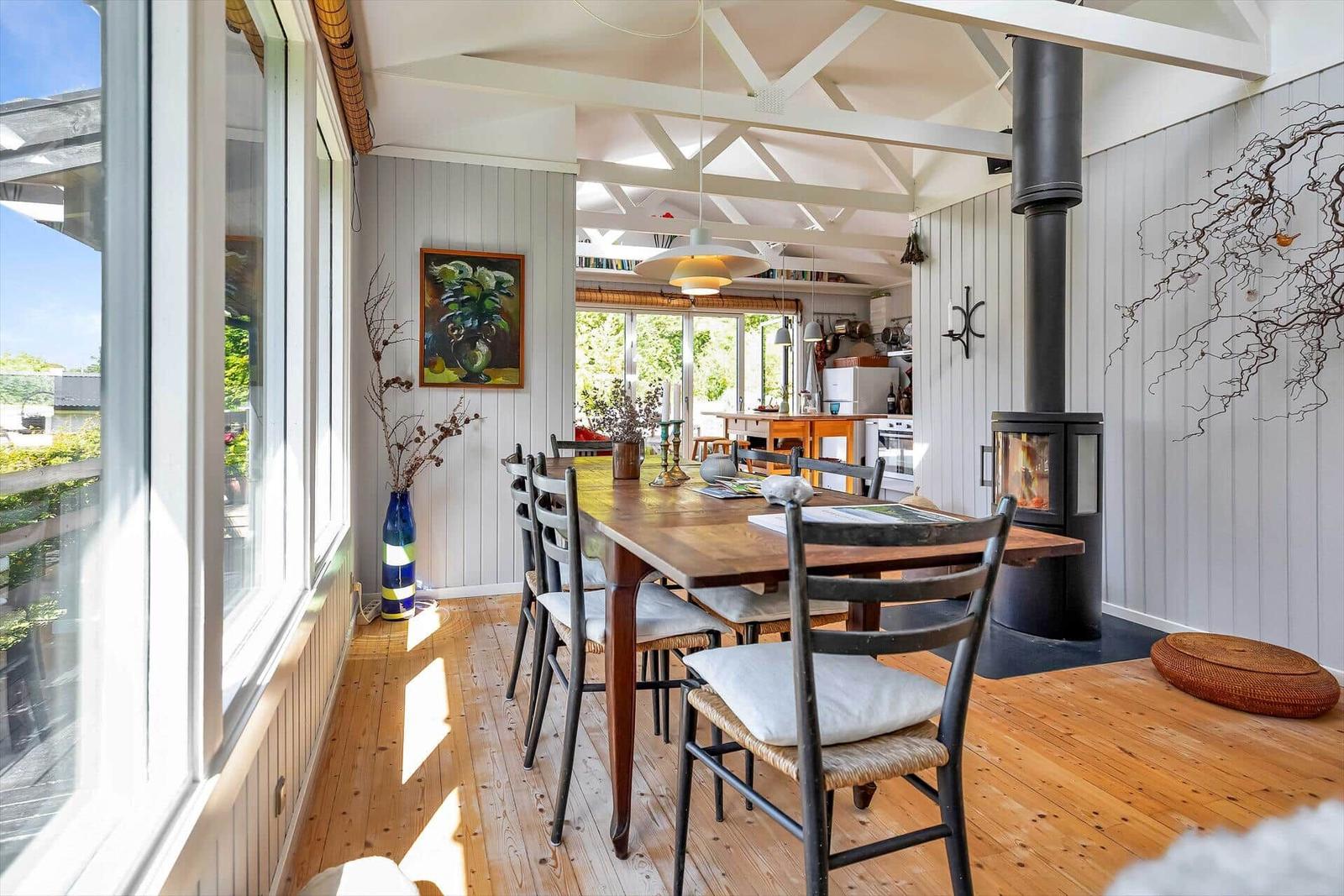 Esszimmer mit Holztisch, Stühlen und Kaminofen. Große Fenster und helles Holzfußboden.