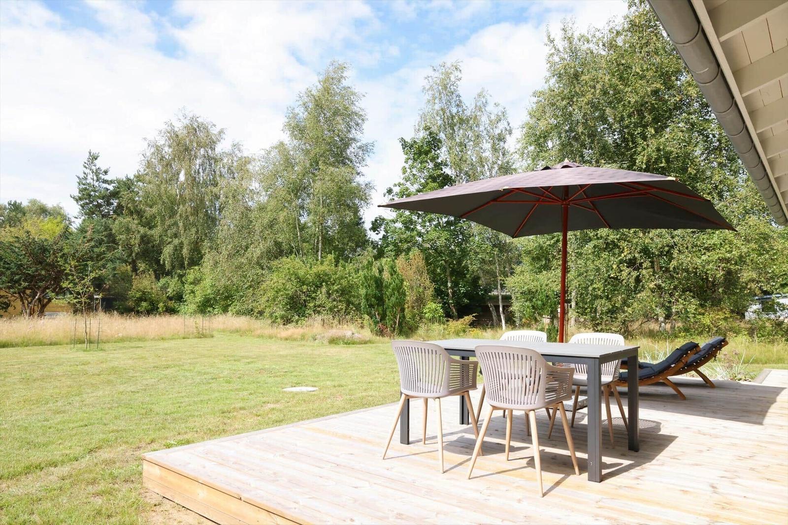 Terrasse mit Tisch, Stühlen und Sonnenschirm. Grasfläche und Bäume im Hintergrund.