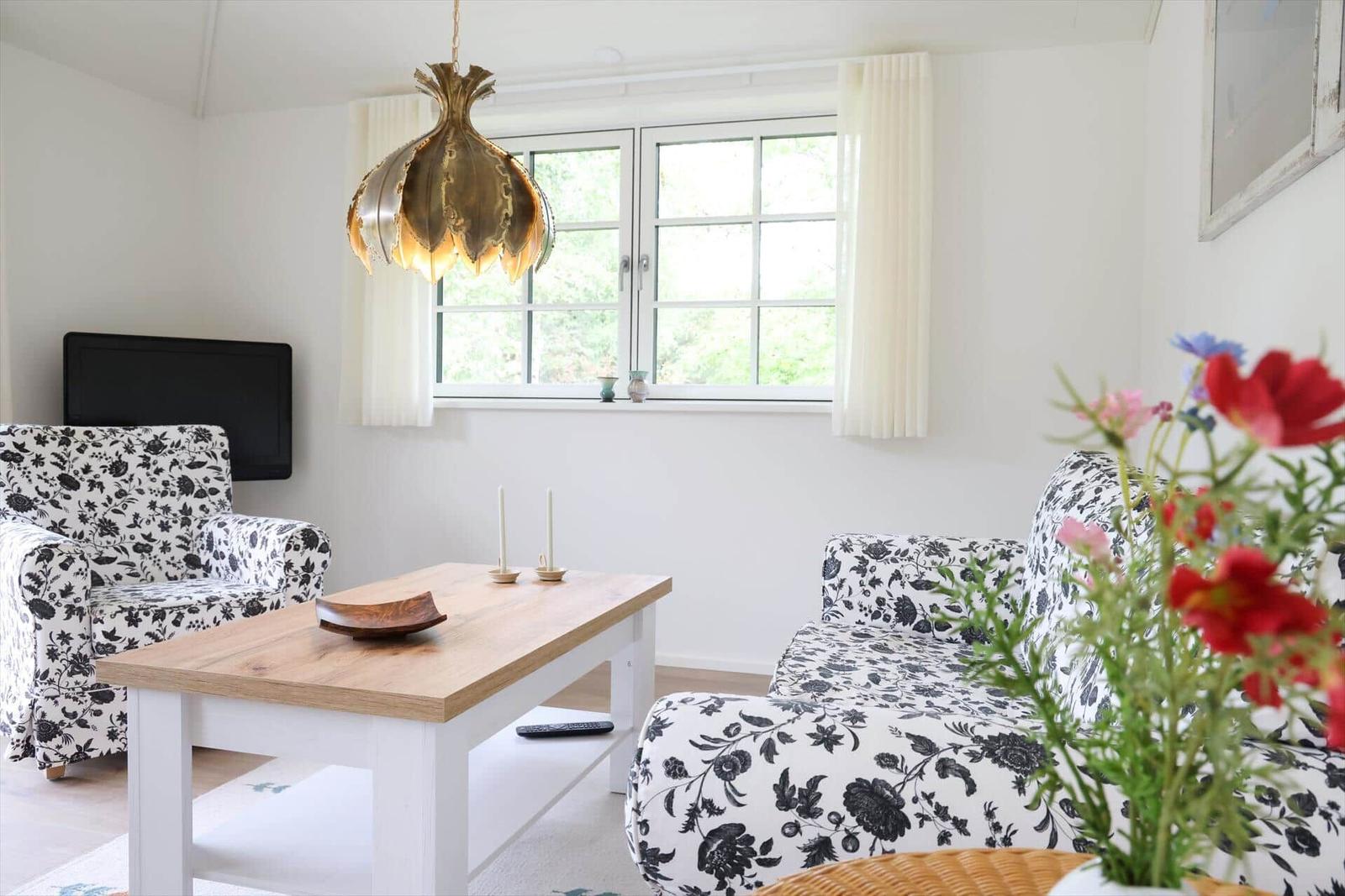 Wohnzimmer mit Sofa, Sessel und Tisch. Fenster mit Vorhängen. Hängelampe und Blumen.