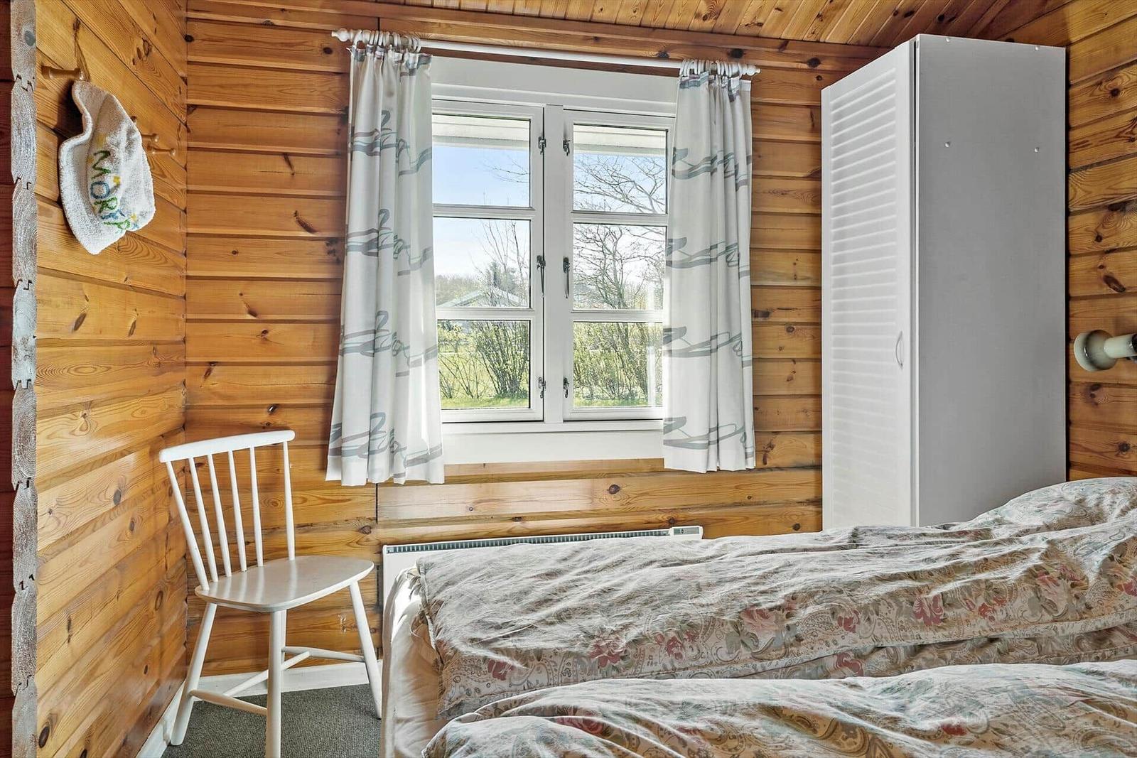 Schlafzimmer mit Holzwand, Fenster, Gardinen, Bett und Stuhl.