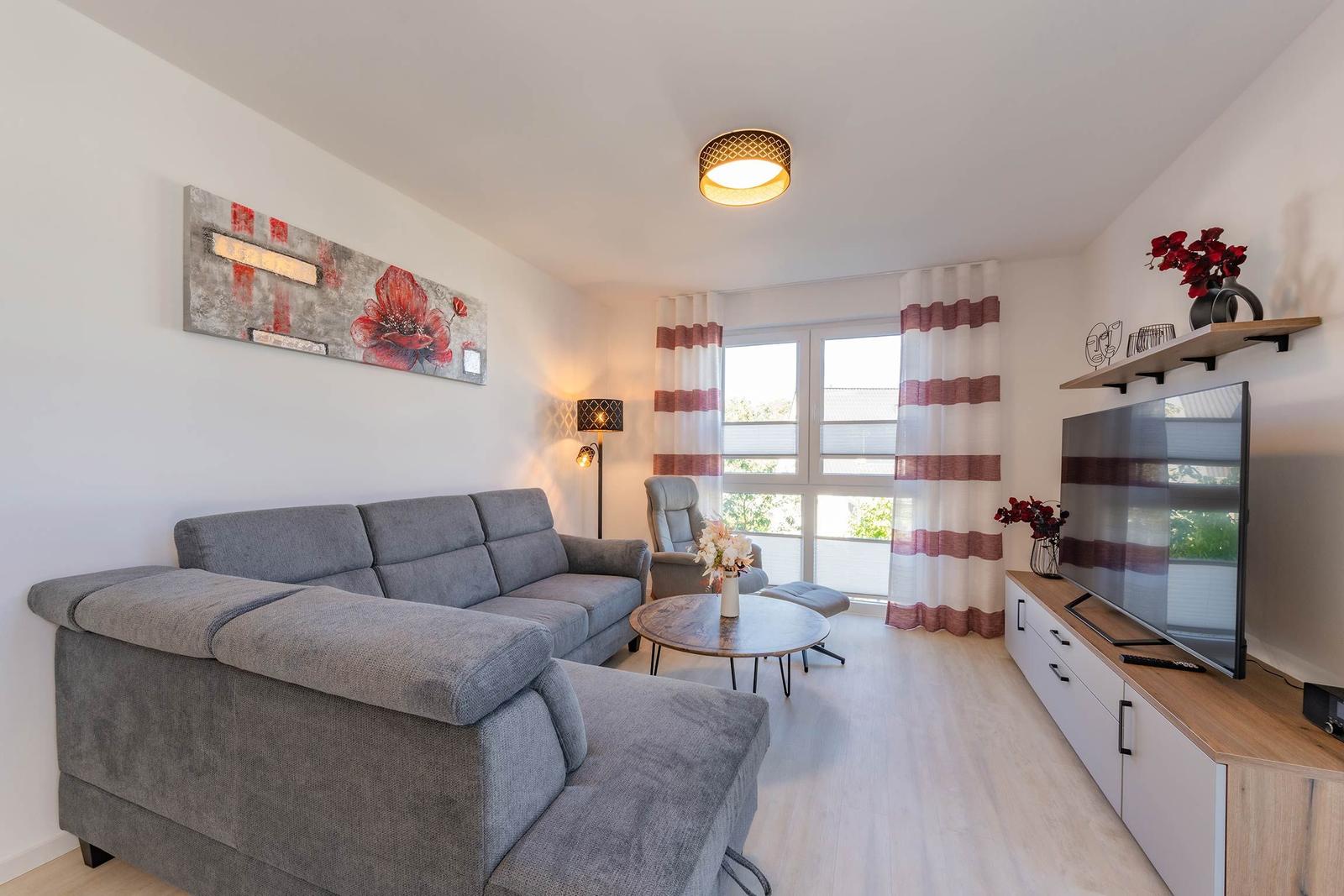 Wohnzimmer mit grauem Sofa, TV und Fenster mit Vorhängen.