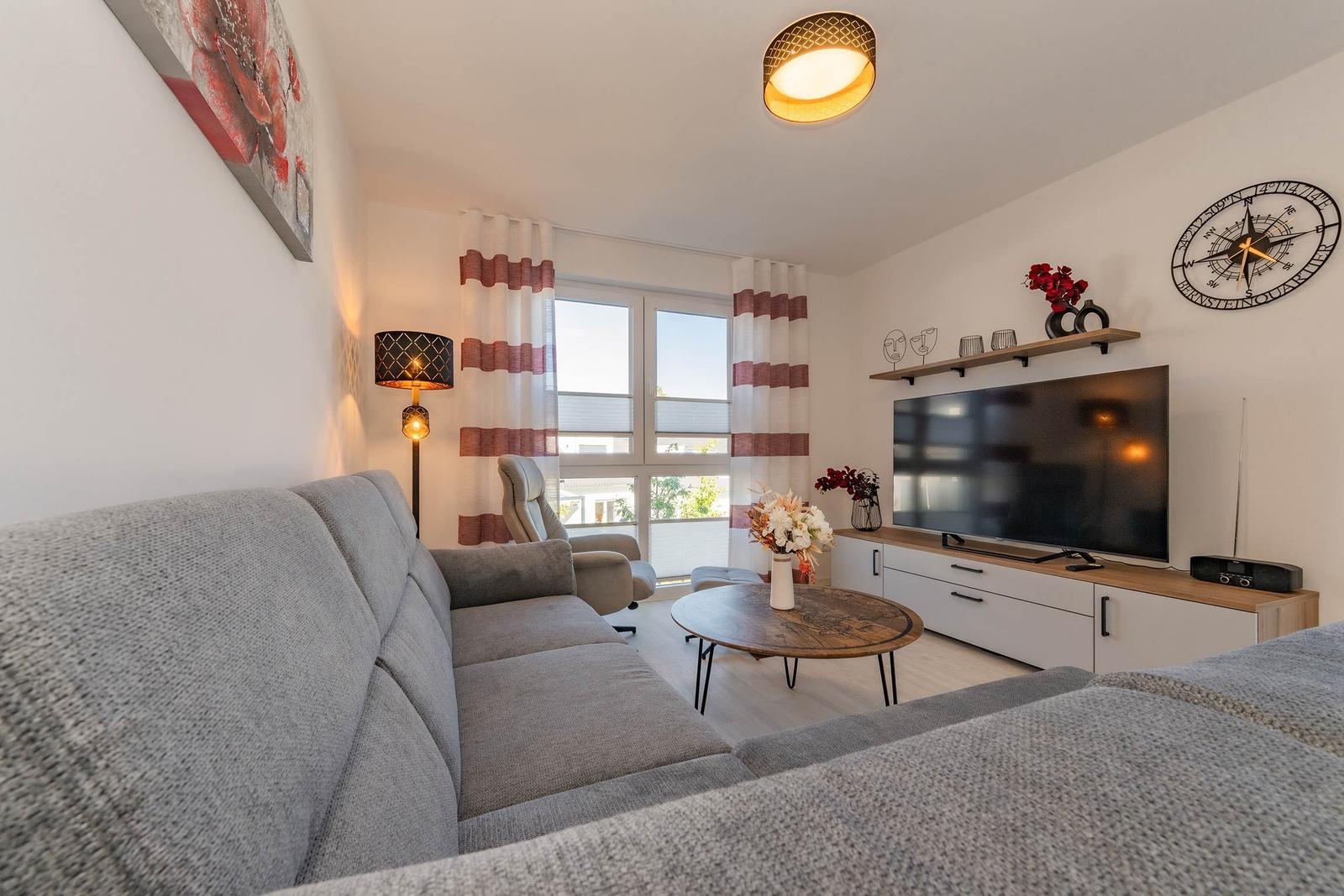 Wohnzimmer mit Sofa, Fernseher und Fenster mit Gardinen.