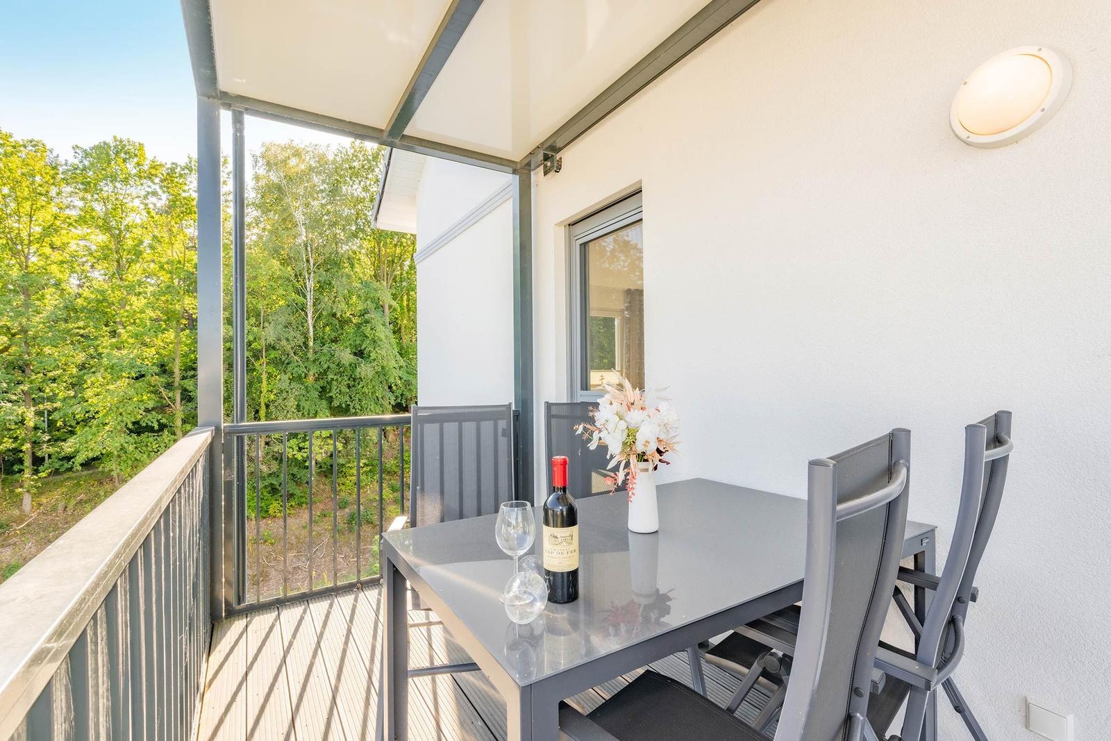 Terrasse mit Tisch, Stühlen, Weinflasche und Blumen. Blick in den Wald.