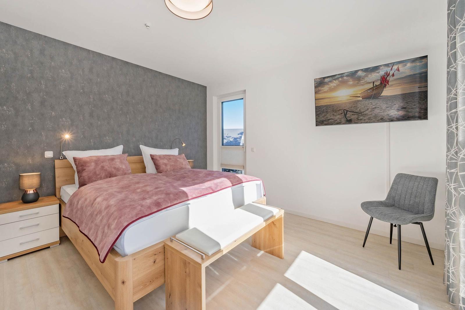 Schlafzimmer mit Bett, Nachttisch und Stuhl. Wandbild und Fenster sichtbar.