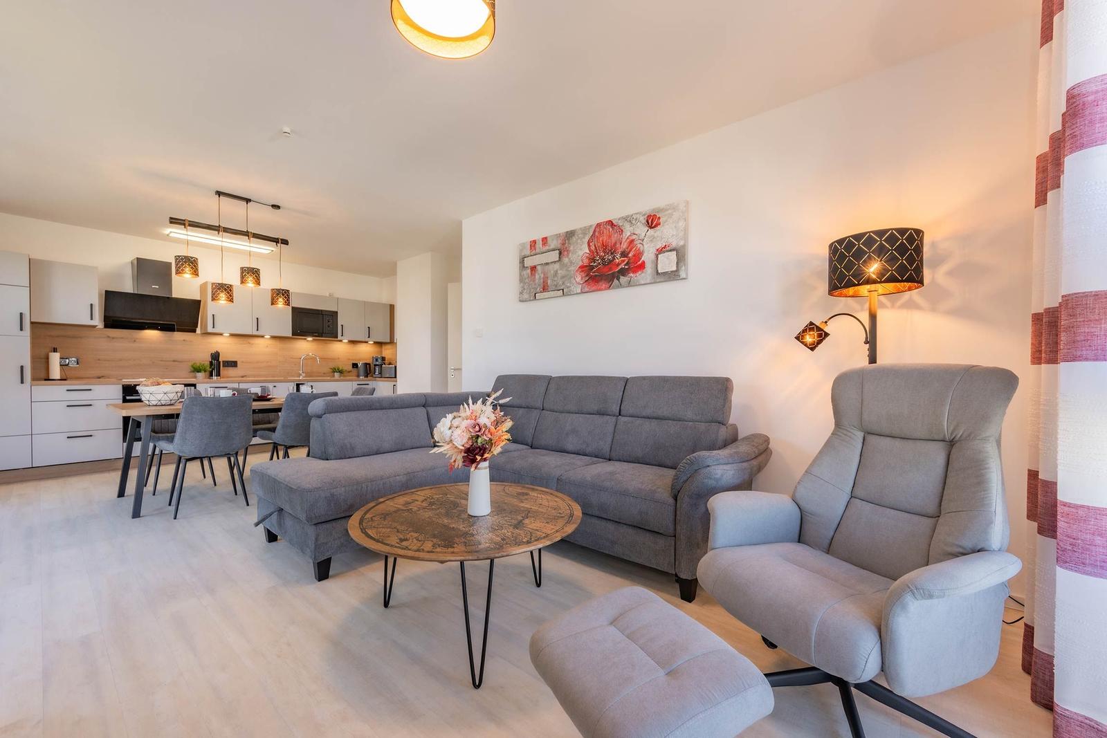 Wohnbereich mit Sofa, Sessel und Küchenbereich im Hintergrund.