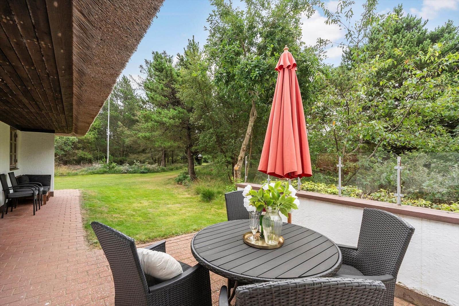Terrasse mit Tisch, Stühlen und geschlossenem Sonnenschirm. Grünfläche und Bäume im Hintergrund.