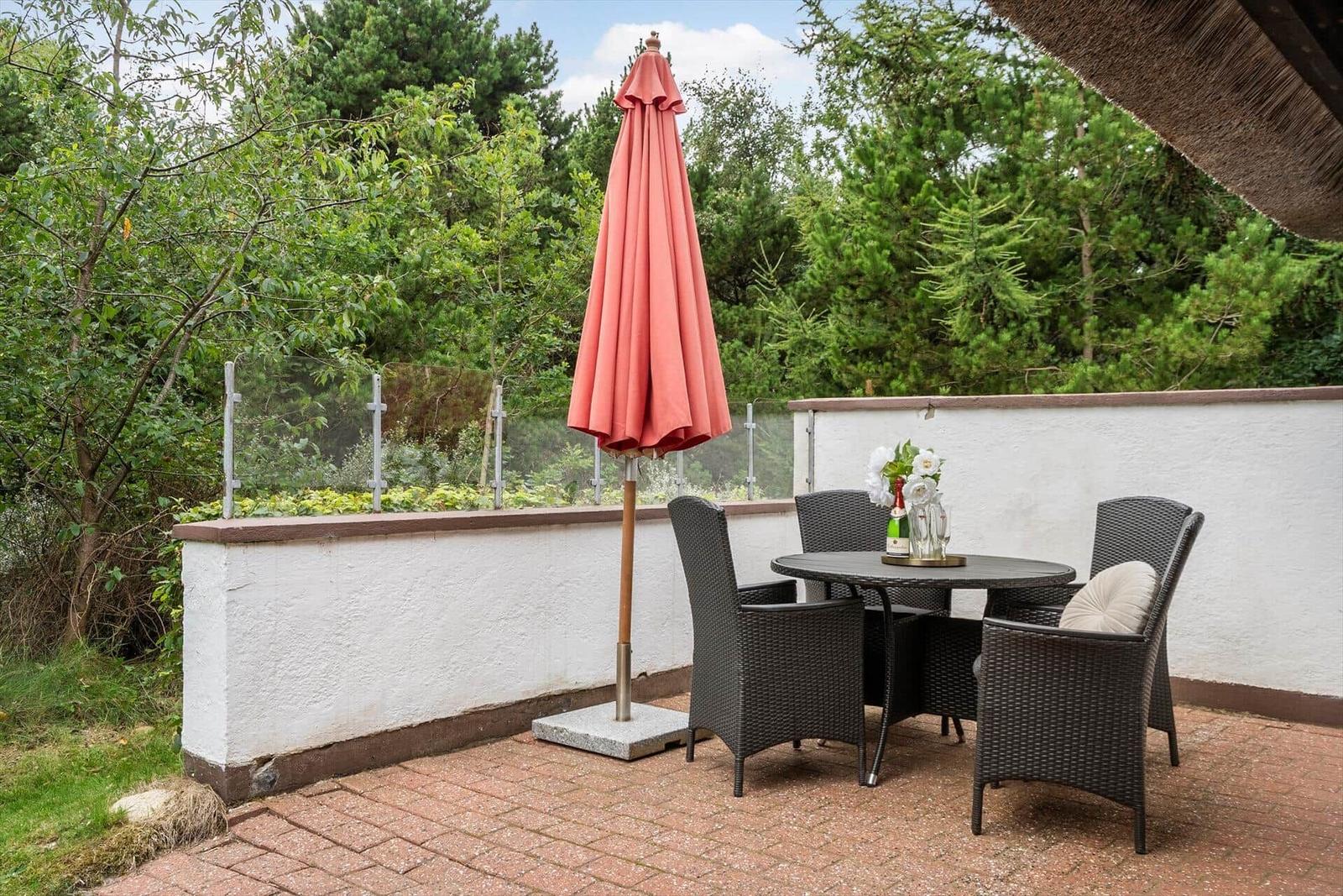 Terrasse mit Tisch, vier Stühlen und geschlossenem Sonnenschirm. Hintergrund: weiße Mauer und Bäume.