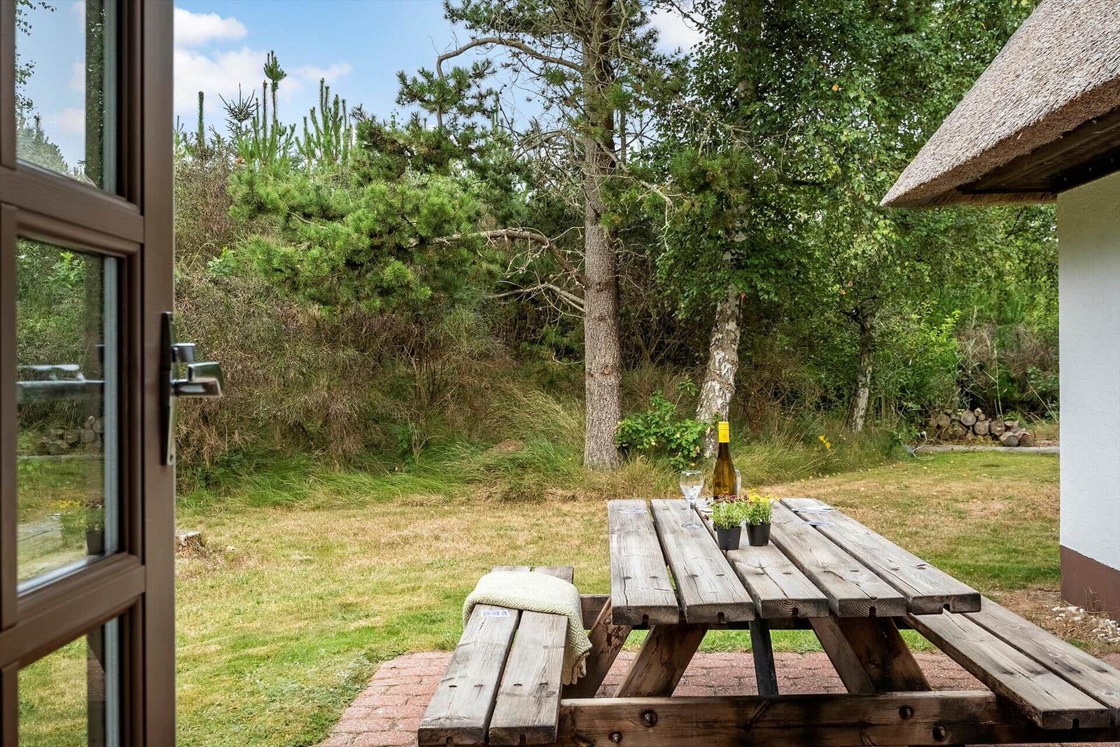 Holztisch mit Flasche und Gläsern auf Terrasse. Grün hinter dem Haus.