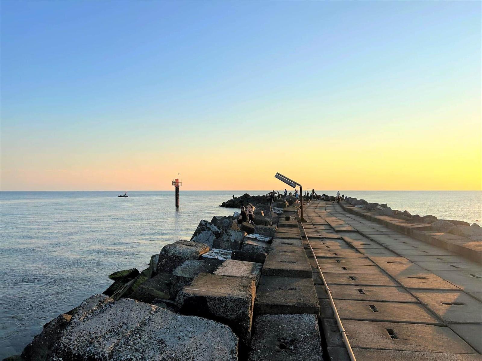 Steinpromenade am Meer bei Sonnenuntergang mit Leuchtturm und Boot.