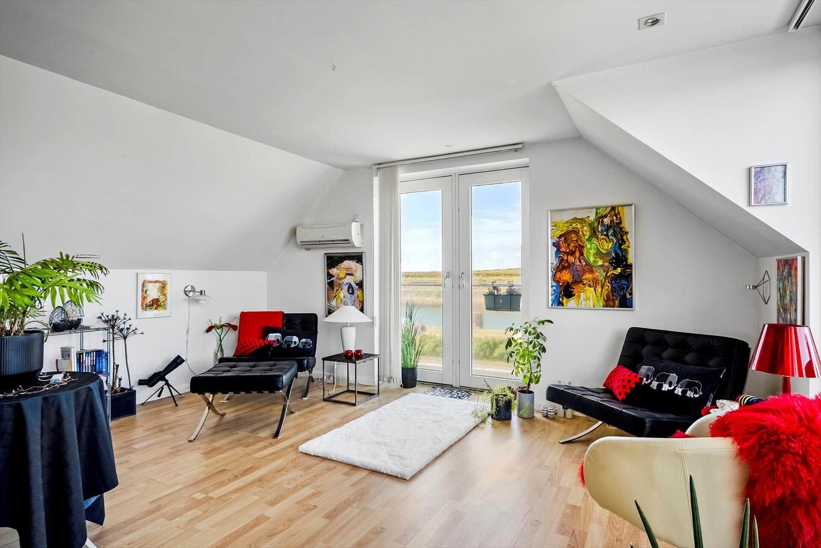 Wohnraum mit Holzboden, schwarzen Sitzmöbeln und roten Akzenten, Blick auf Wasser.