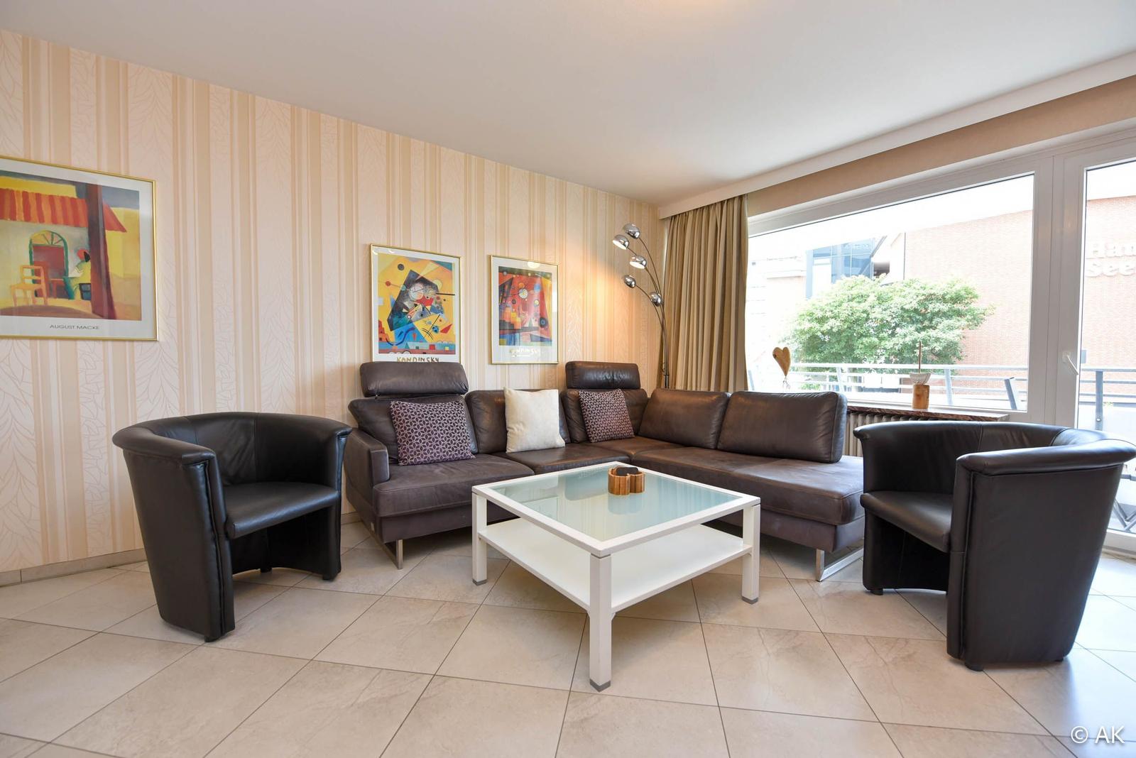 Wohnzimmer mit braunem Sofa, zwei Sesseln und Glas-Tisch. Wand mit Tapete und Kunstwerken.