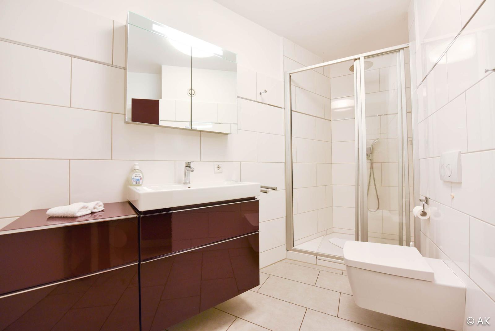 Badezimmer mit Dusche, Waschbecken und Toilette. Fliesenwand und dunkler Schrank.