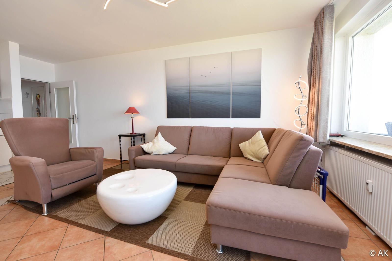 Wohnzimmer mit Eckcouch, Sessel und runden Tisch. Wandbild und Fenster sichtbar.