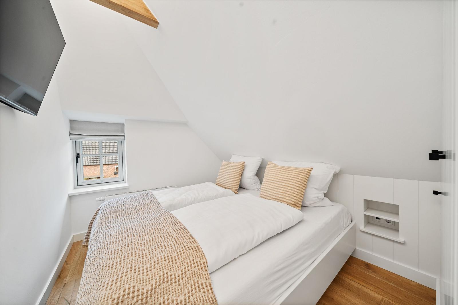 Schlafzimmer mit Dachschräge, Bett, Fenster und Fernseher an der Wand.