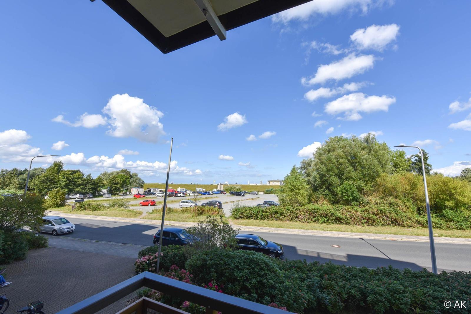 Blick von Balkon auf Straße, Parkplatz und grüne Bäume unter blauem Himmel mit weißen Wolken.