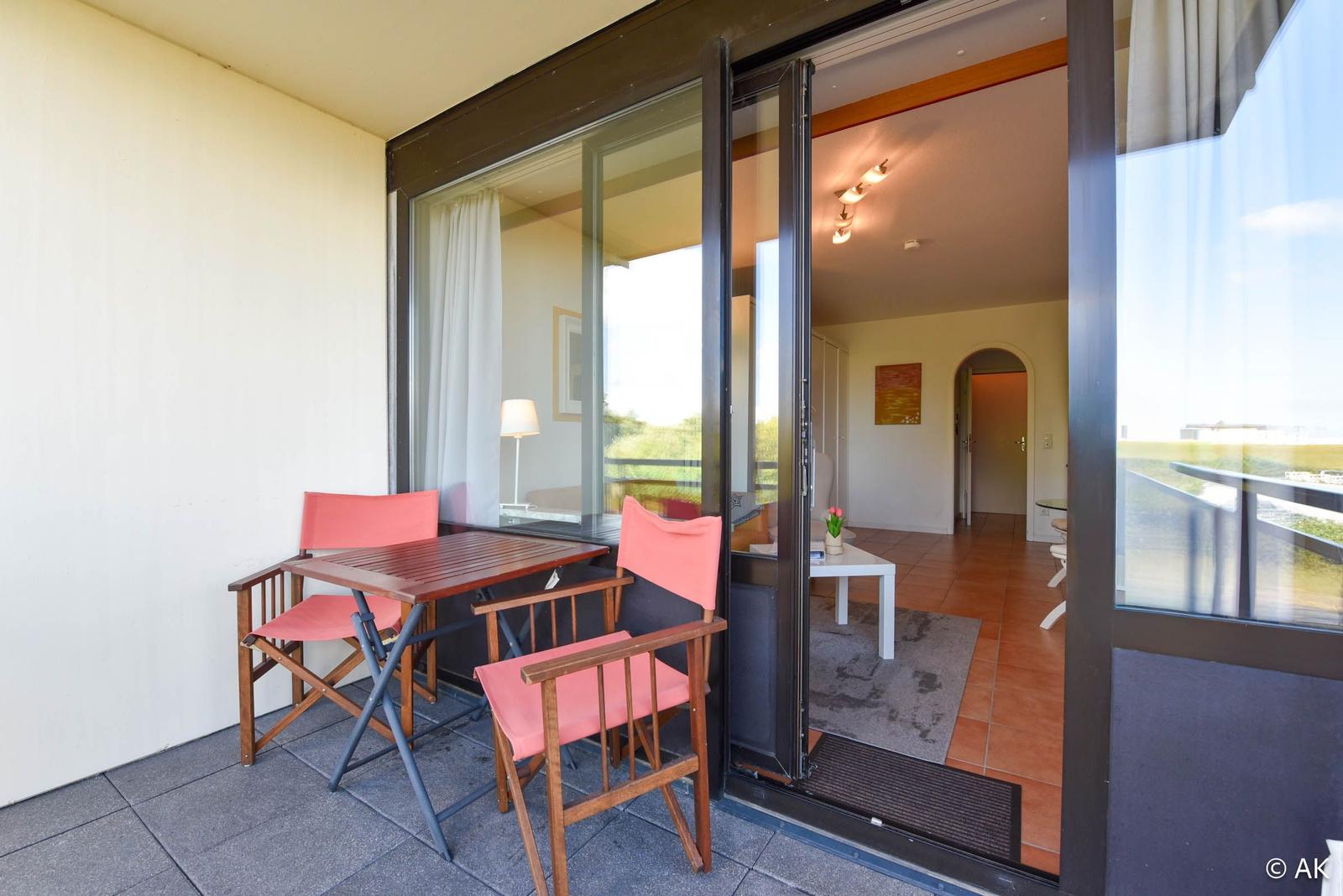 Terrasse mit Tisch und zwei Stühlen. Durch Glastür ist Wohnbereich sichtbar.