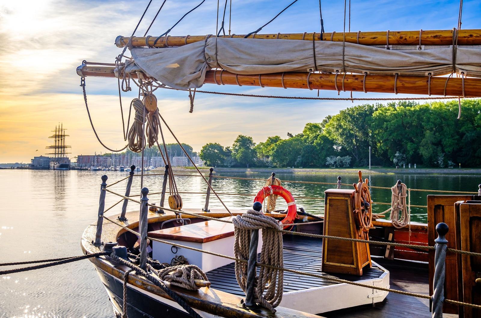Vorderdeck eines Segelschiffs mit Ruder und Rettungsring am Wasser.