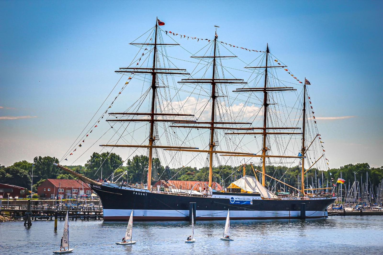 Großes Segelschiff mit mehreren Masten liegt im Hafen. Kleine Segelboote sind im Wasser.