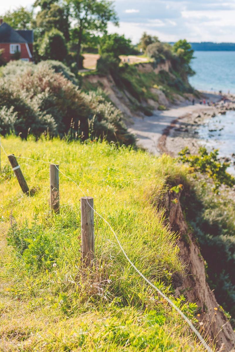 Grünes Gras mit Holzpfosten und Drahtzaun an steilem Hang über Strand und Meer.