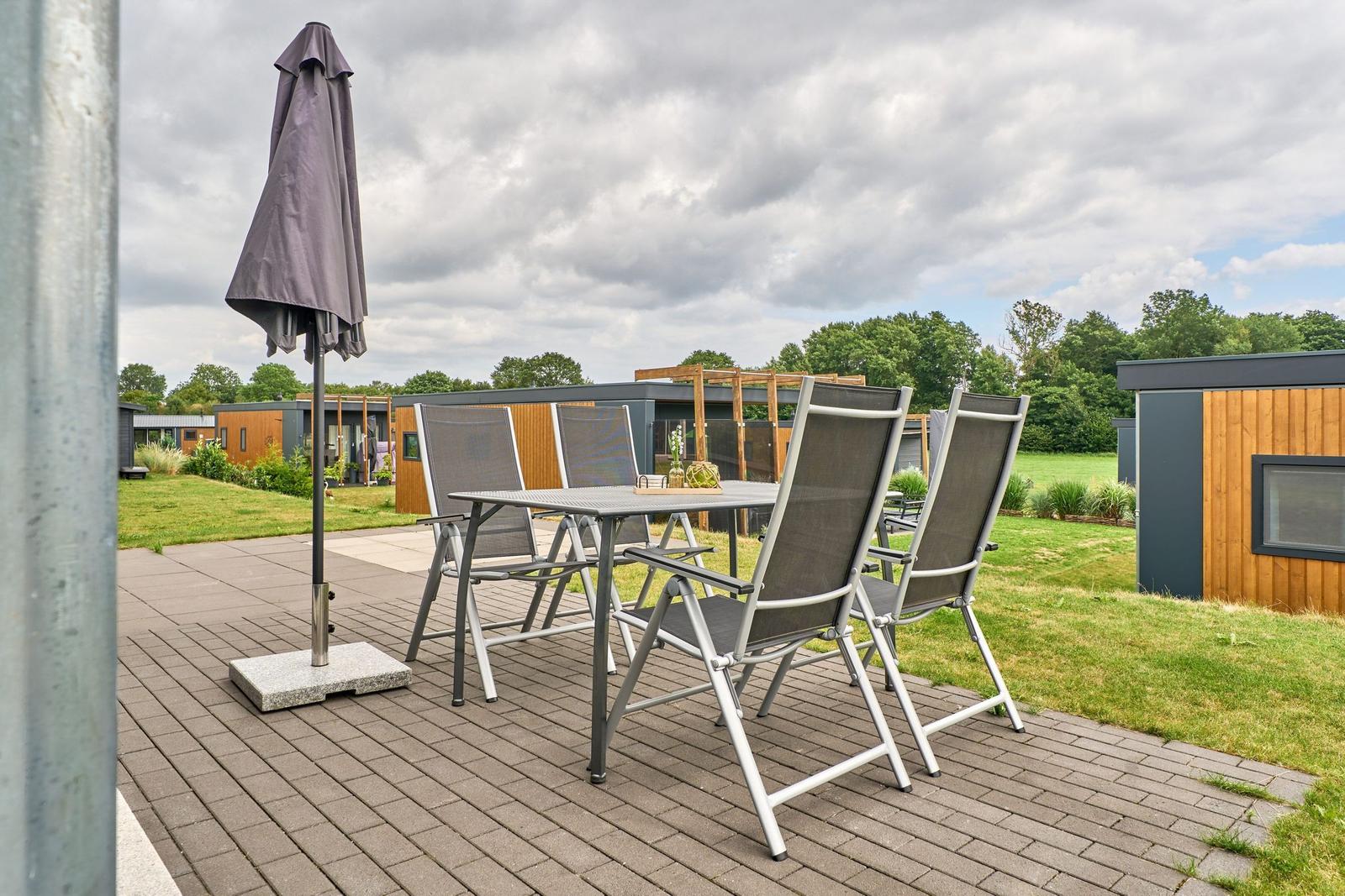 Terrasse mit Tisch, vier Stühlen und geschlossenem Sonnenschirm. Hintergrund: moderne Häuser und Grünfläche.