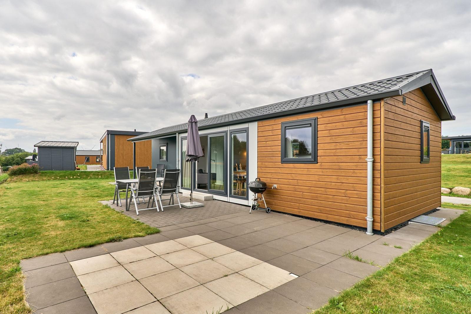 Moderne Holzhütte mit Terrasse, Grill und Garten. Glastüren führen ins Innere.