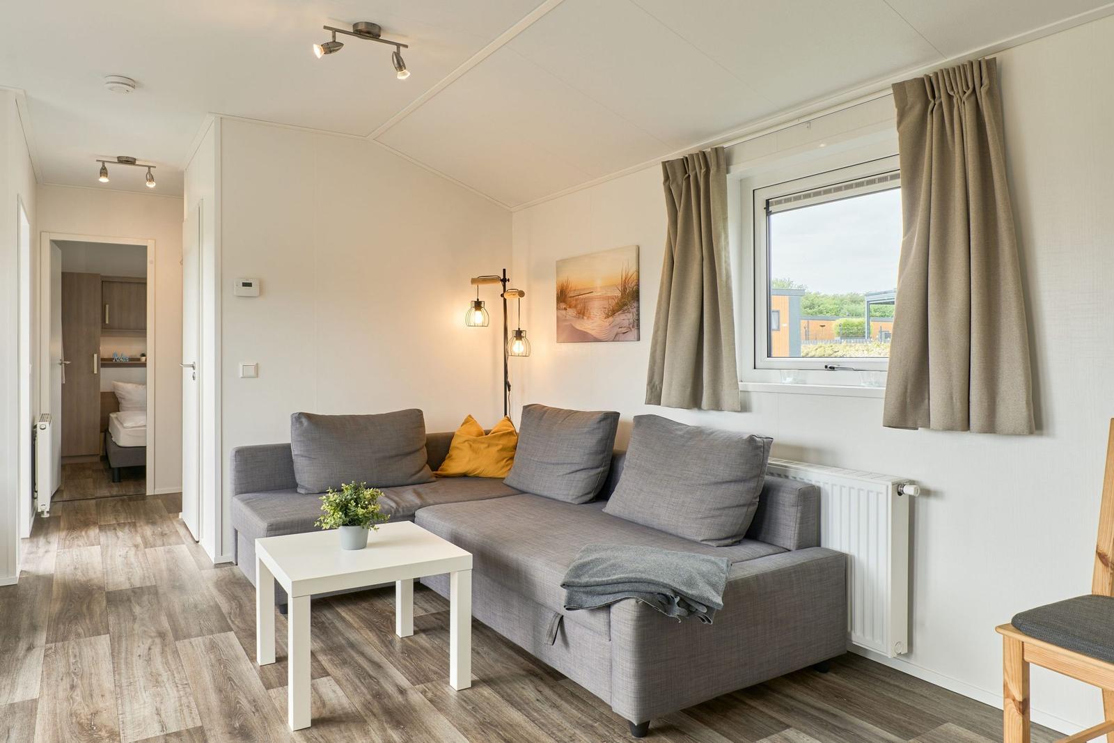 Wohnzimmer mit Sofa, Tisch und Fenster. Im Hintergrund ist das Schlafzimmer zu sehen.