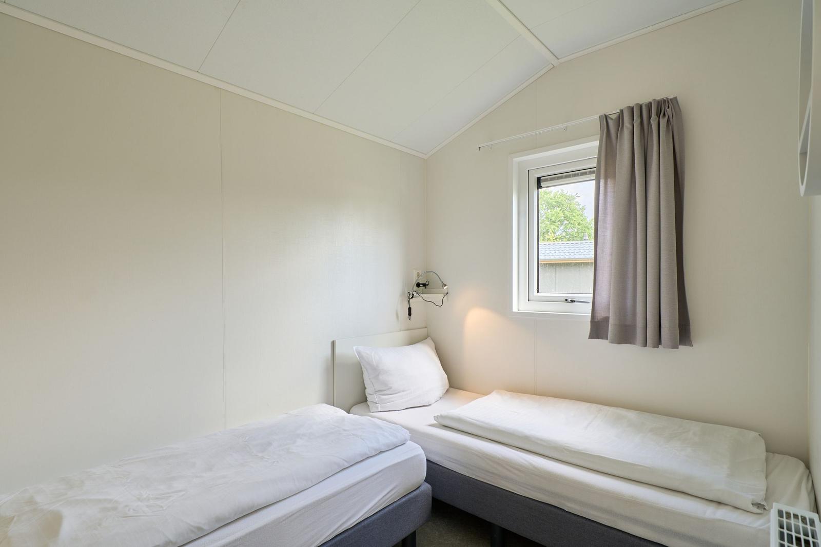 Zwei Einzelbetten mit weißen Bettwäschen, Fenster mit Vorhang und Wandlampe.